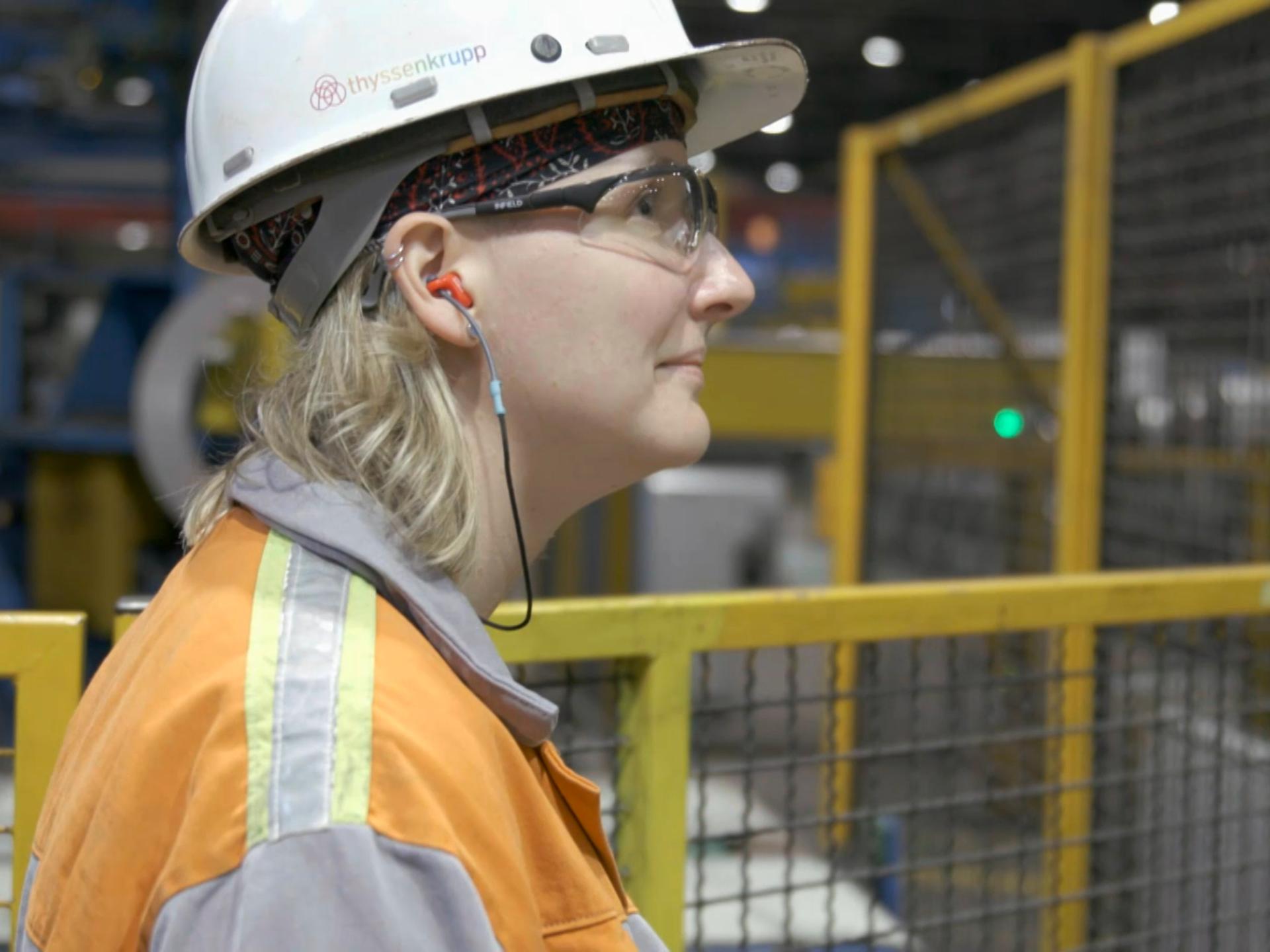 Ann Catherine Meyer steht mit Schutzhelm und Arbeitsjacke in einer Produktionshalle von Thyssenkrupp und blickt nach rechts 