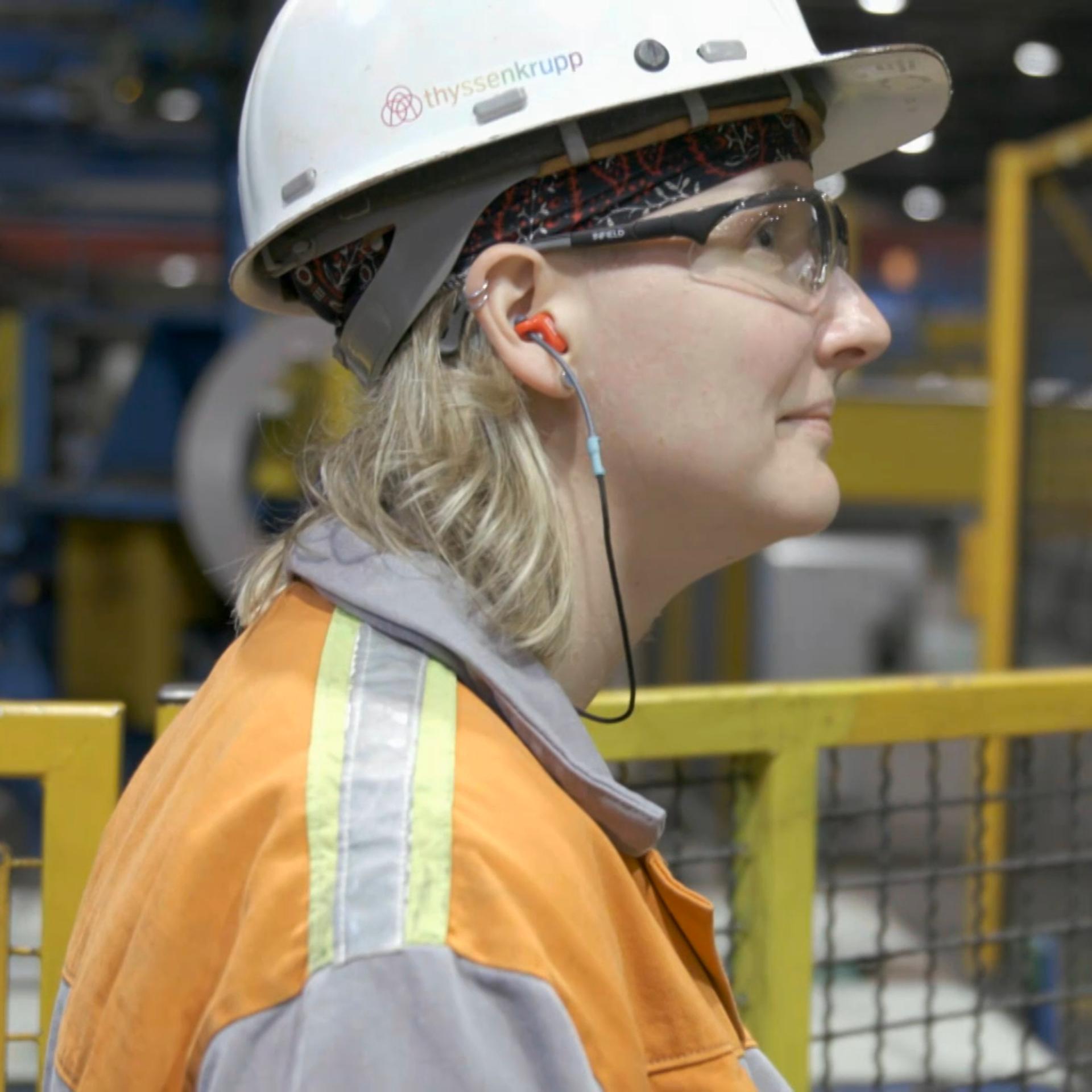 Ann Catherine Meyer steht mit Schutzhelm und Arbeitsjacke in einer Produktionshalle von Thyssenkrupp und blickt nach rechts 