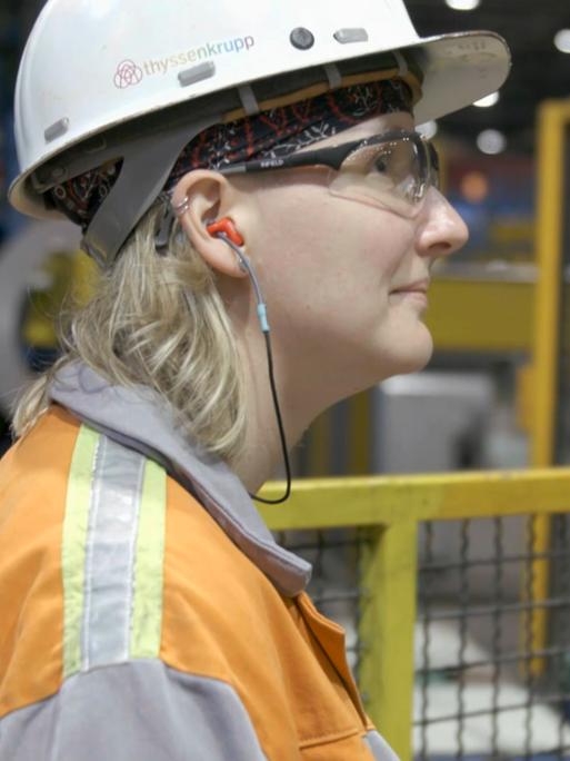 Ann Catherine Meyer steht mit Schutzhelm und Arbeitsjacke in einer Produktionshalle von Thyssenkrupp und blickt nach rechts Ann Catherine Meyer steht mit Schutzhelm und Arbeitsjacke in einer Produktionshalle von Thyssenkrupp und blickt nach rechts