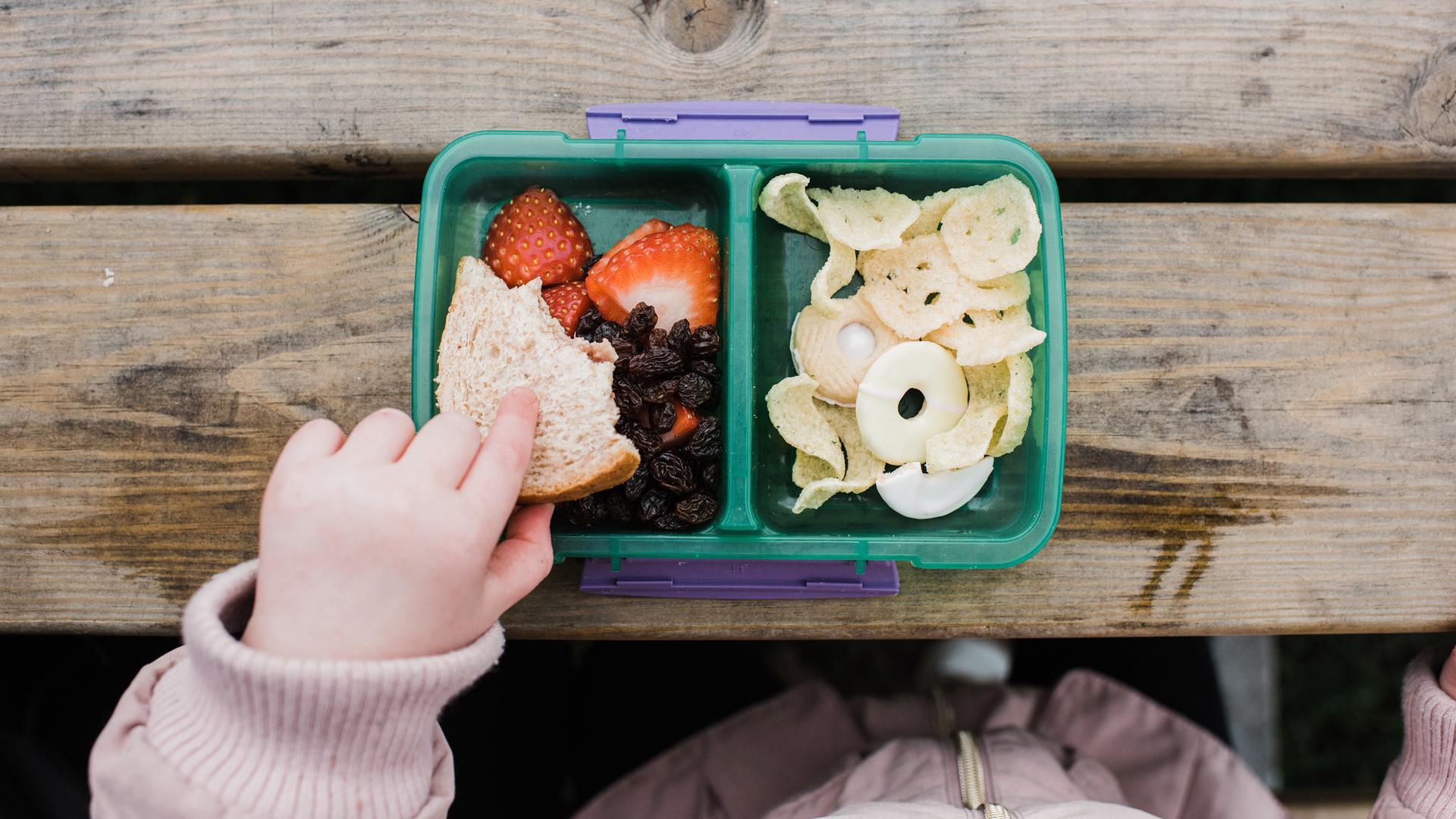 Ein kleines Kinder isst mit der Hand Obststückchen aus einer Lunchbox auf einem Holztisch.
