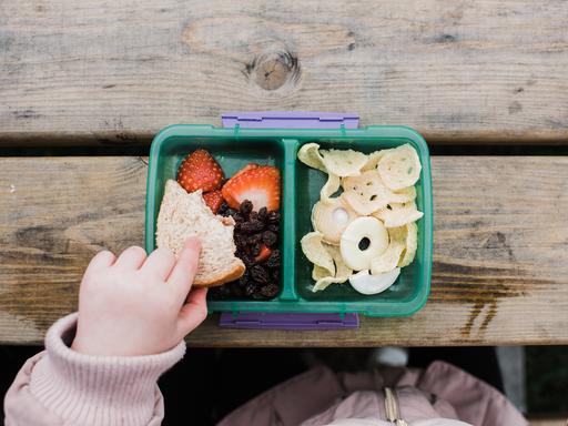 Ein kleines Kinder isst mit der Hand Obststückchen aus einer Lunchbox auf einem Holztisch.