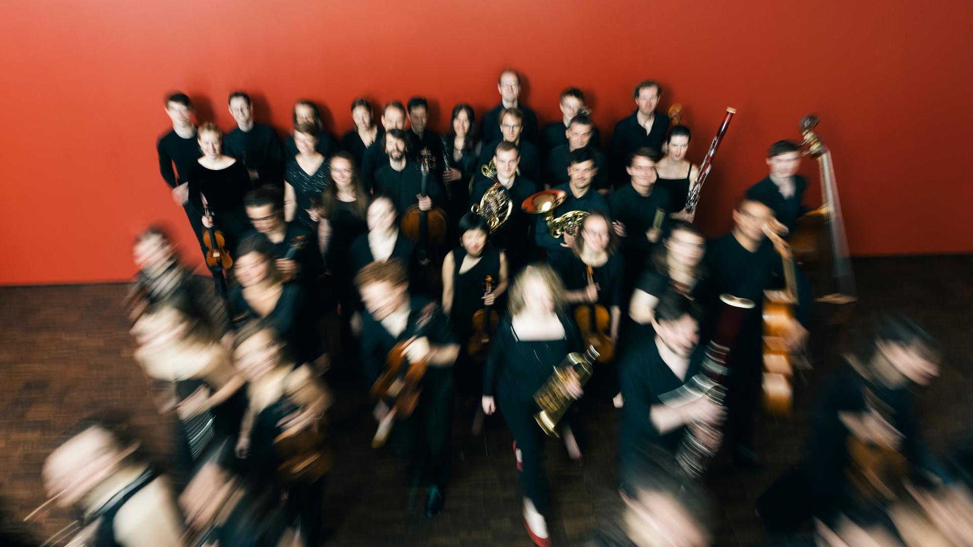Das Ensemble reflektor steht in einem Raum mit roter Wand, aus dem sie sich entfernen. Durch den Kameraeffekt ist ihre Bewegung durch verschwommene Personen deutlich gemacht. Das Ensemble reflektor steht in einem Raum mit roter Wand, aus dem sie sich entfernen. Durch den Kameraeffekt ist ihre Bewegung durch verschwommene Personen deutlich gemacht.