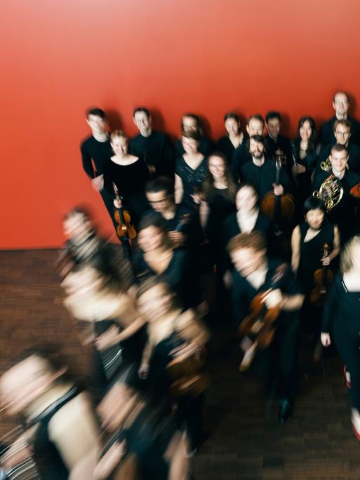 Das Ensemble reflektor steht in einem Raum mit roter Wand, aus dem sie sich entfernen. Durch den Kameraeffekt ist ihre Bewegung durch verschwommene Personen deutlich gemacht.