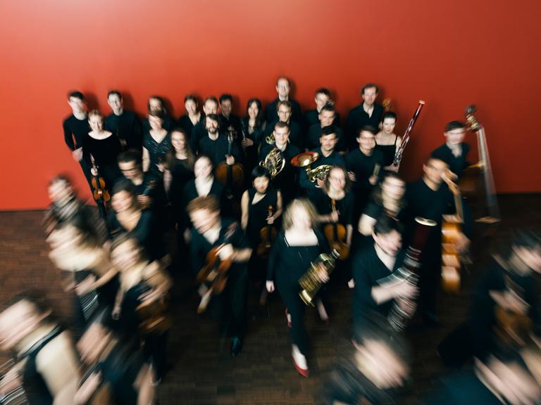Das Ensemble reflektor steht in einem Raum mit roter Wand, aus dem sie sich entfernen. Durch den Kameraeffekt ist ihre Bewegung durch verschwommene Personen deutlich gemacht.
