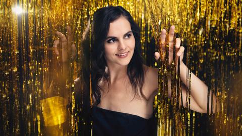 Pianistin Danae Dörken greift mit den Fingern auf Augenhöhe in die Fäden eines goldenen Fadenvorhanges, durch den sie gerade mit ihren offenen, schwarzen Haaren hindurchtritt.