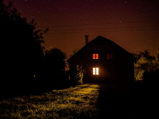 Nachts: Blick auf ein einsames Haus auf einer Wiese, am Rand Bäume.