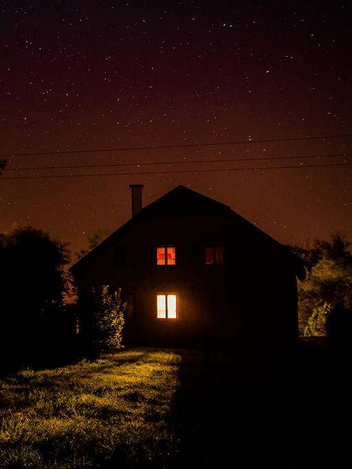 Nachts: Blick auf ein einsames Haus auf einer Wiese, am Rand Bäume.