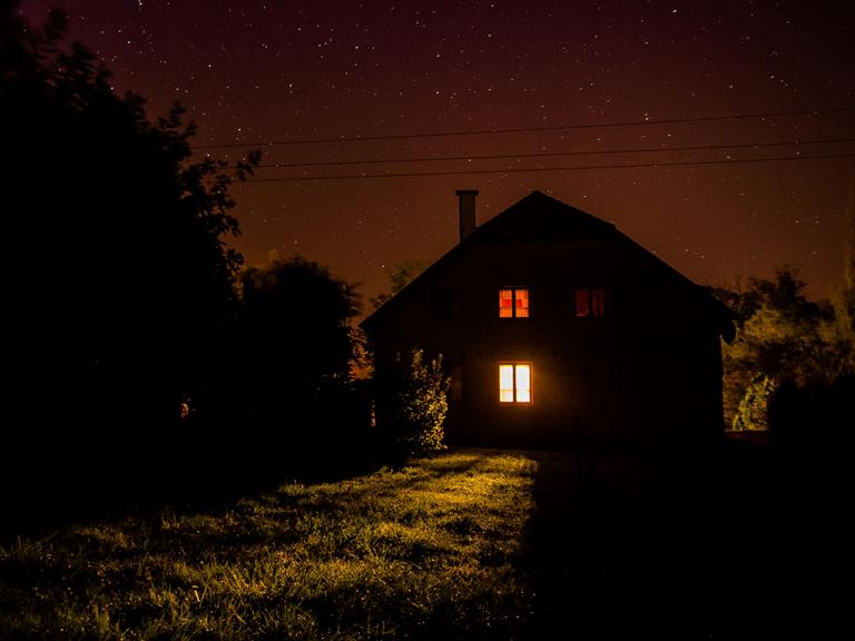 Nachts: Blick auf ein einsames Haus auf einer Wiese, am Rand Bäume.