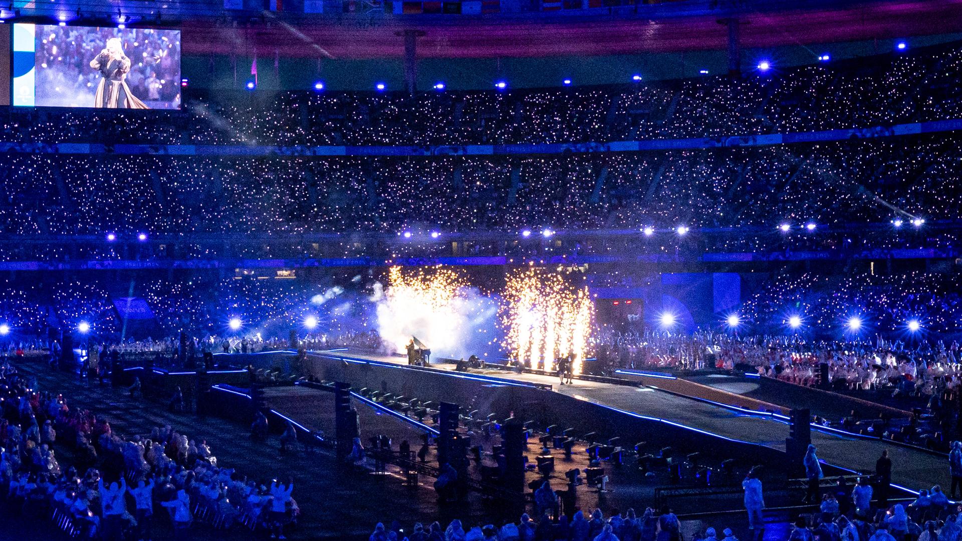 Eindrücke von der Abschlussfeier der Paralympischen Spiele: Das vollbesetzte Stade de France ist in blaues Licht getaucht. In der Mitte ist eine Bühne mit einem Feuerwerk.