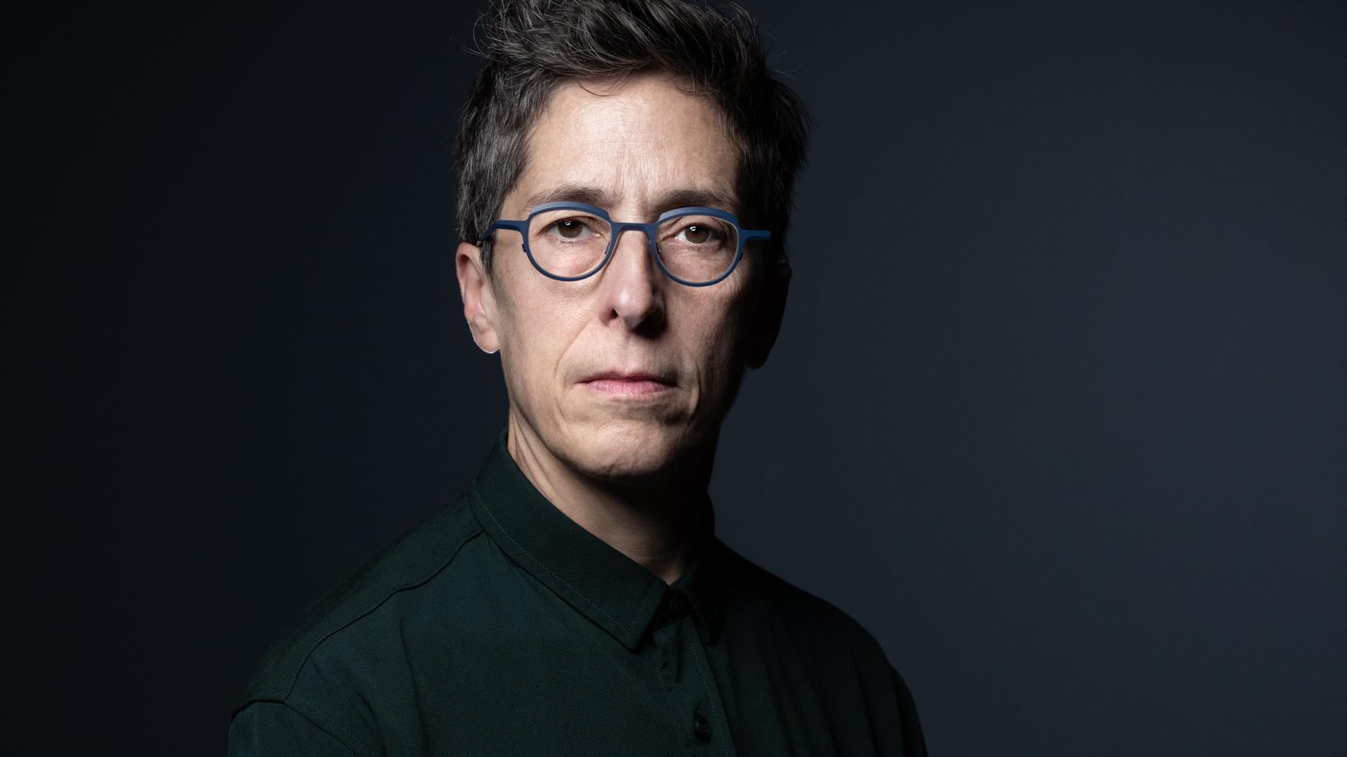 Alison Bechdel sitzt vor einem schwarzen Hintergrund und schaut direkt in die Kamera. Sie hat kurzes, dunkles Haar und trägt eine grau umrandete Brille.
