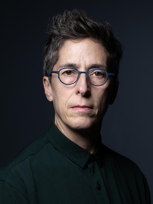 Alison Bechdel sitzt vor einem schwarzen Hintergrund und schaut direkt in die Kamera. Sie hat kurzes, dunkles Haar und trägt eine grau umrandete Brille.