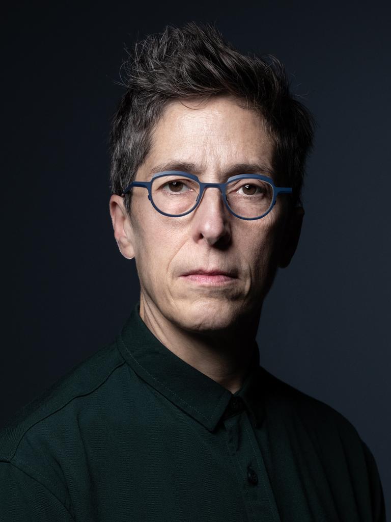 Alison Bechdel sitzt vor einem schwarzen Hintergrund und schaut direkt in die Kamera. Sie hat kurzes, dunkles Haar und trägt eine grau umrandete Brille.