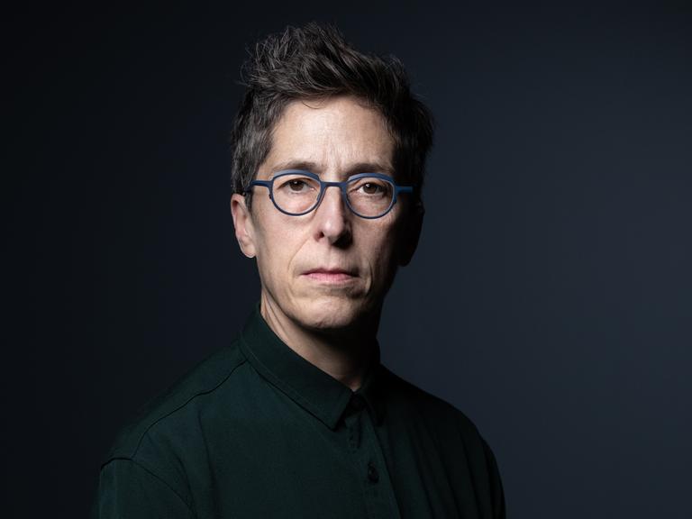 Alison Bechdel sitzt vor einem schwarzen Hintergrund und schaut direkt in die Kamera. Sie hat kurzes, dunkles Haar und trägt eine grau umrandete Brille.