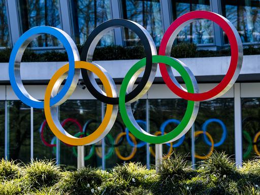 Die olympischen Ringe in den Farben Blau, Schwarz, Rot, Gelb und Grün sind vor einem Gebäude angebracht.