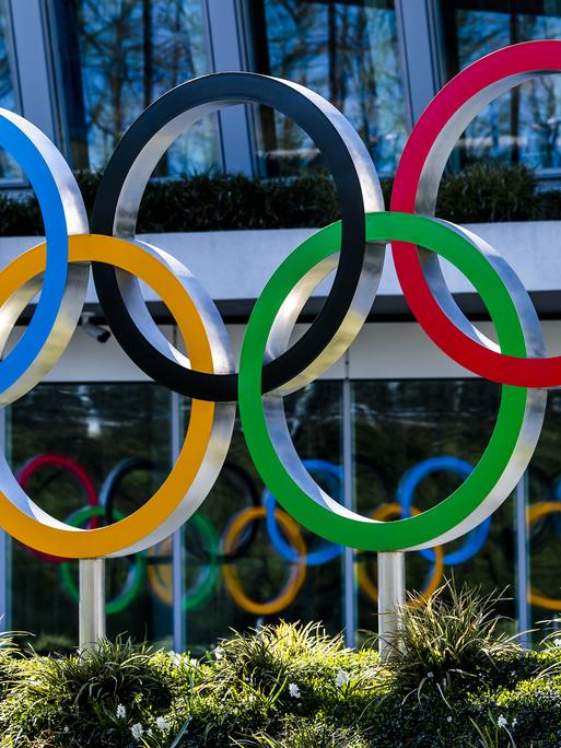 Die olympischen Ringe in den Farben Blau, Schwarz, Rot, Gelb und Grün sind vor einem Gebäude angebracht.