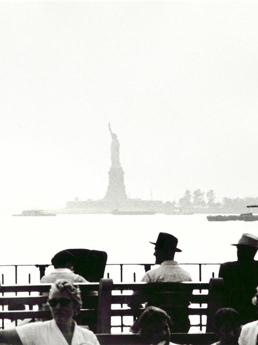 Menschen sitzen in New York am Ufer mit Blick auf die Freiheitsstatue.