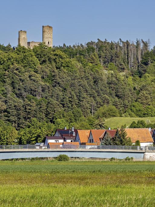Blick auf eine Burgruine über einem Dorf mit Brücke, umgeben von grünen Wäldern und Wiesen unter klarem Himmel. Die Ruine Brandenburg im Werratal bei Lauchröden in Thüringen (Symbolbild) 