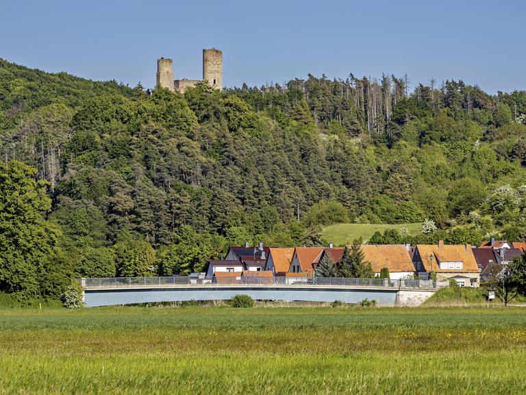 Blick auf eine Burgruine über einem Dorf mit Brücke, umgeben von grünen Wäldern und Wiesen unter klarem Himmel. Die Ruine Brandenburg im Werratal bei Lauchröden in Thüringen (Symbolbild) 