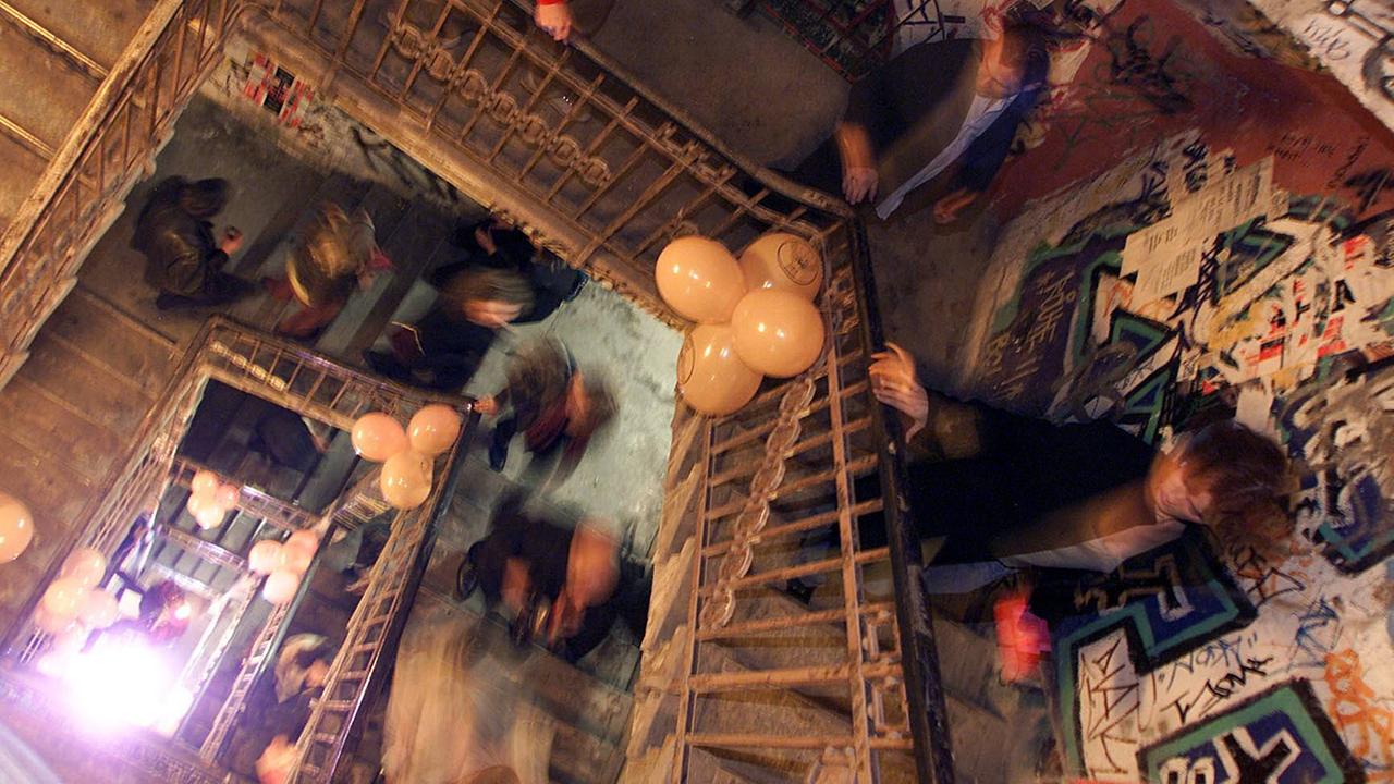 Menschen laufen eine Treppe nach oben, am Geländer hängen Luftballons, die Wänder sind voller Graffiti.