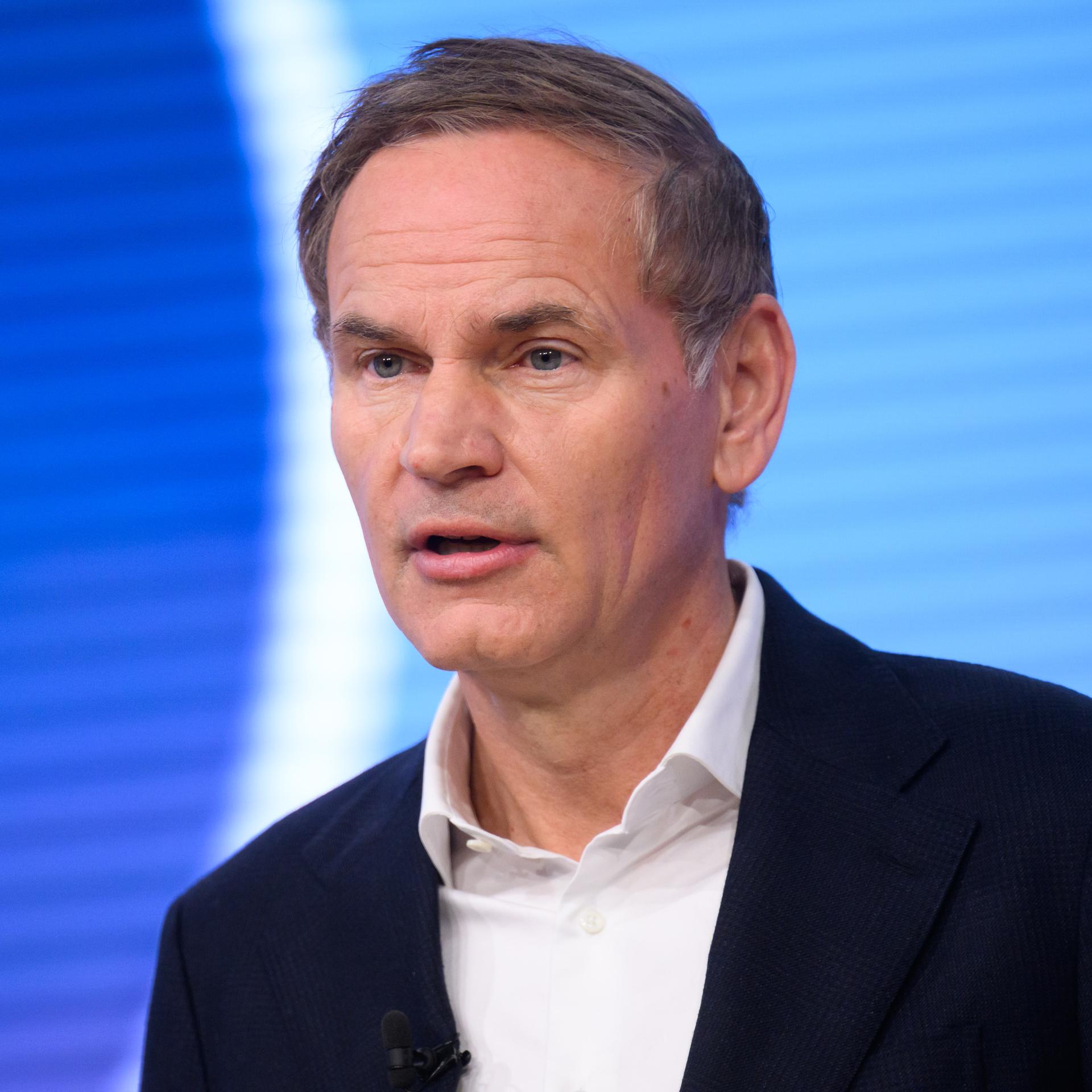 Blume steht vor einer blauen LED-Wand mit weißen Streifen und sagte etwas. Er trägt ein dunkles jackett und ein weißes Hemd. 