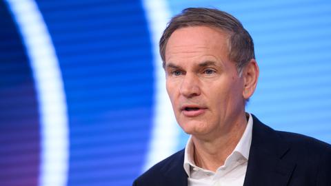 Blume steht vor einer blauen LED-Wand mit weißen Streifen und sagte etwas. Er trägt ein dunkles jackett und ein weißes Hemd. 