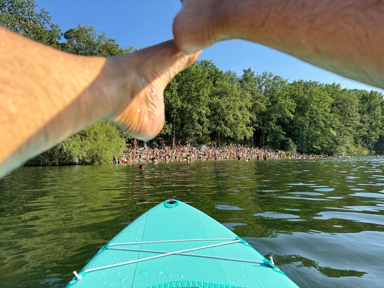 Menschen am Ufer des Sees Krumme Lanke in Berlin aus der Perspektive einer Person auf dem Wasser auf einem Standup-Board.