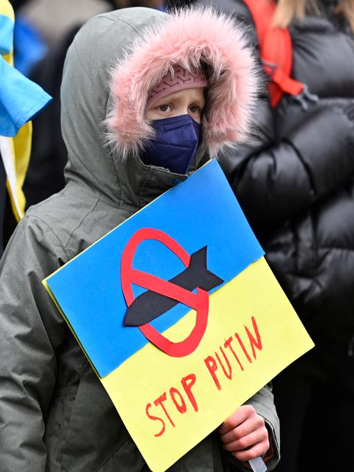 Ein junger Demonstrant hält ein Plakat in den Farben der ukrainischen Flagge, mit der Aufschrift „Stop Putin“ und der Darstellung einer Rakete, die mit einem roten Verbotszeichen versehen ist, in seinen Händen.