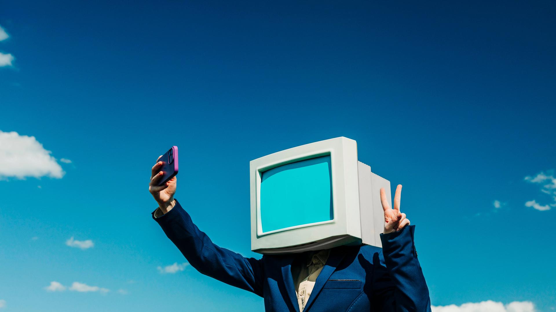 Eine Person in einem blauen Anzug trägt auf den Schultern einen Computerbildschirm, es ist eine Attrappe. Die Person macht ein Selfie von sich mit einem Smartphone.