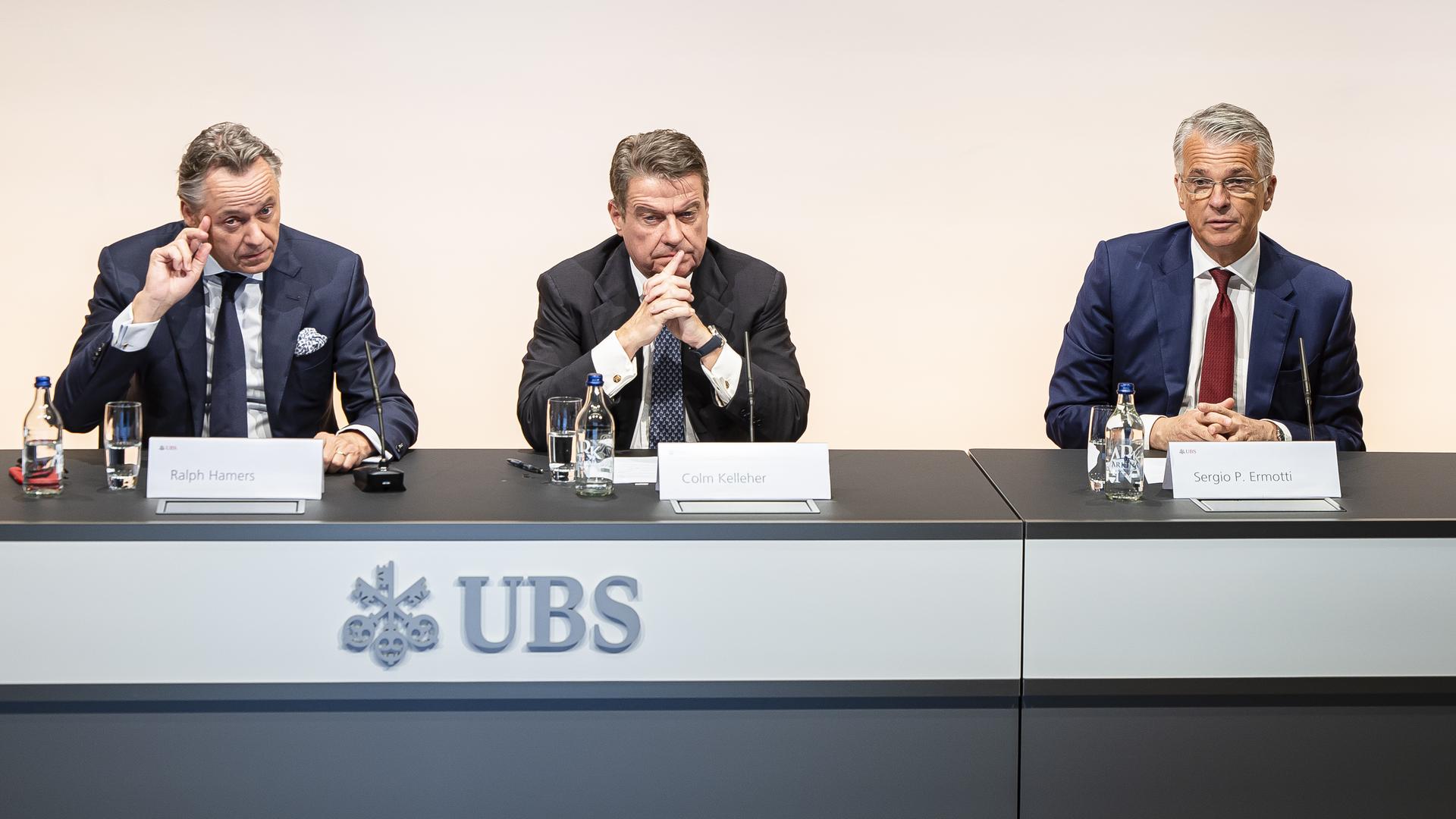 Drei Männer in Anzügen während einer Pressekonferenz, auf ihrem Tisch steht das Logo UBS