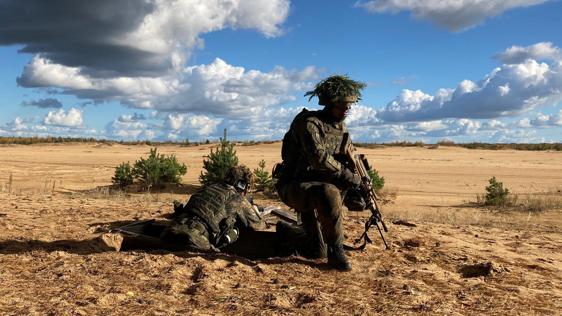  Ein getarnter Soldat kniet auf trockenem, braunen Boden und hält ein Gewehr mit dem Lauf nach unten.