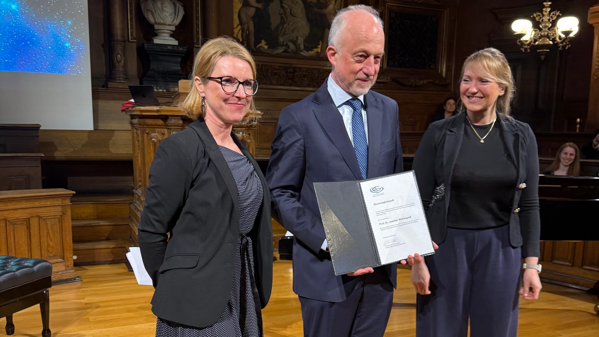 Joachim Wambsganß steht mit der der AG-Ehrenurkunde in den Händen zwischen Janine Fohlmeister (links) und Stefanie Walch-Gassner, beide im Vorstand der Deutschen Astronomischen Gesellschaft. Joachim Wambsganß steht mit der der AG-Ehrenurkunde in den Händen zwischen Janine Fohlmeister (links) und Stefanie Walch-Gassner, beide im Vorstand der Deutschen Astronomischen Gesellschaft.