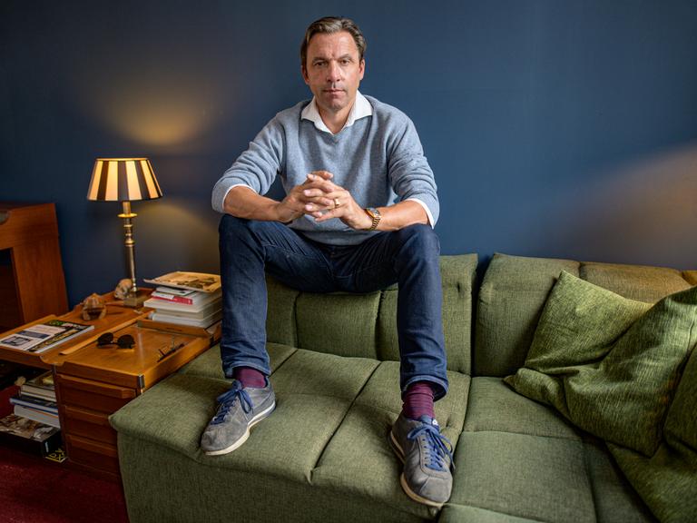 Rocko Schamoni sitzt auf der Lehne eines grünen Sofas, während seine Schuhe auf der Sitzfläche ruhen. Dabei schaut er verschmitzt in die Kamera.