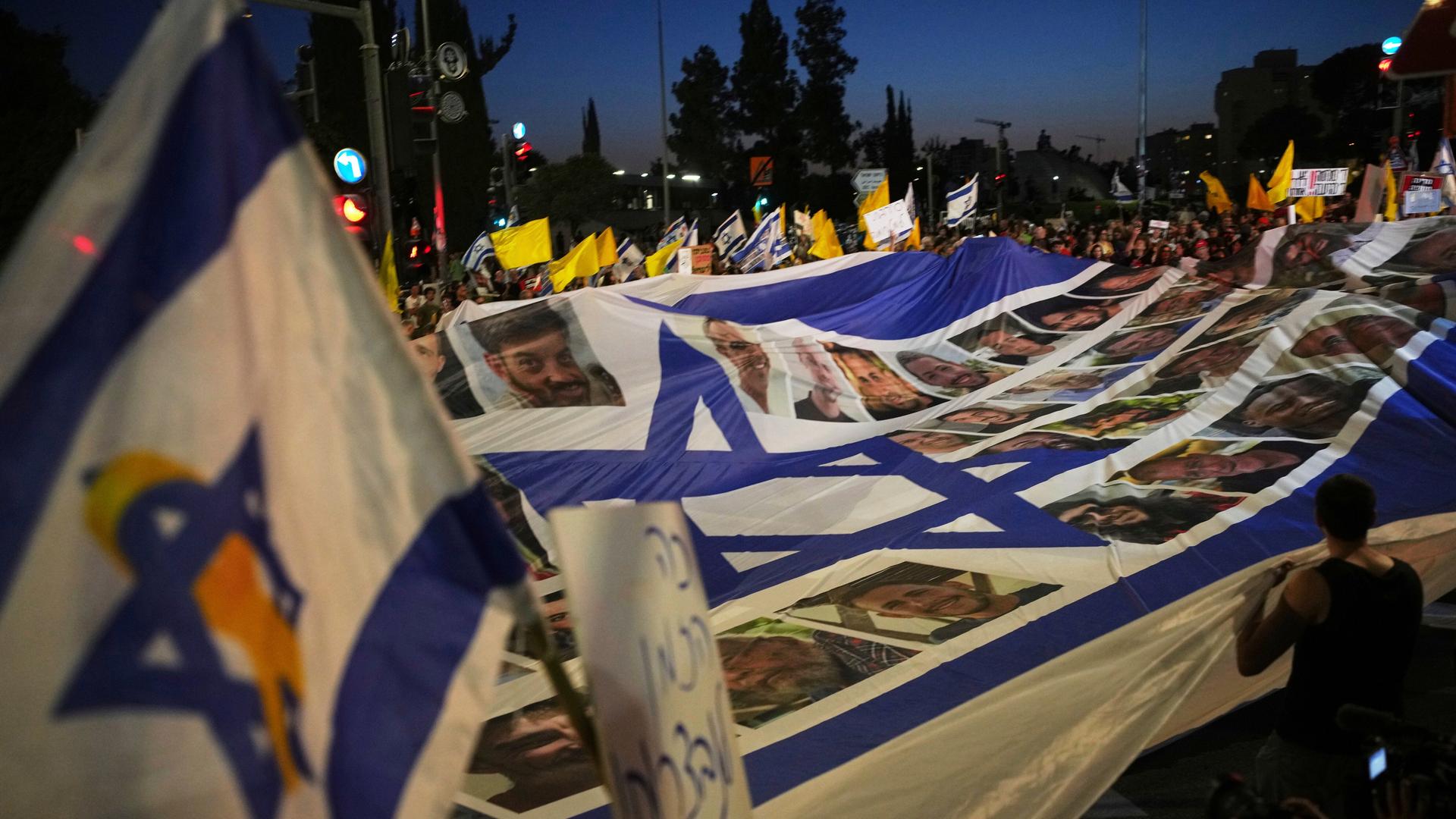 Eine große israelische Flagge trägt Bilder von Geiseln, drumherum stehen Menschen