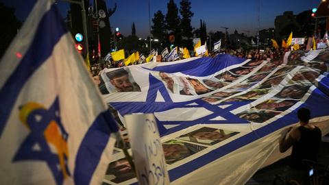 Eine große israelische Flagge trägt Bilder von Geiseln, drumherum stehen Menschen