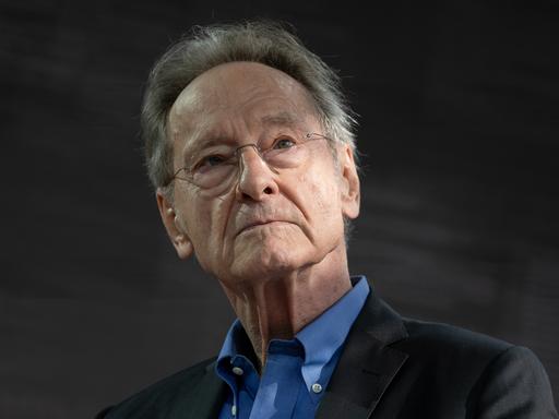 Ein Mann mit grauen Haaren, Brille und schwarzem Sakko schaut in Richtung der Kamera.