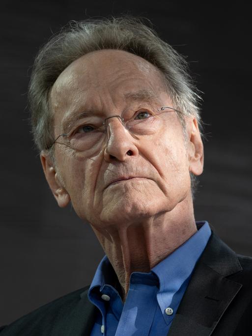 Ein Mann mit grauen Haaren, Brille und schwarzem Sakko schaut in Richtung der Kamera.