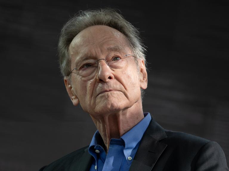 Ein Mann mit grauen Haaren, Brille und schwarzem Sakko schaut in Richtung der Kamera.