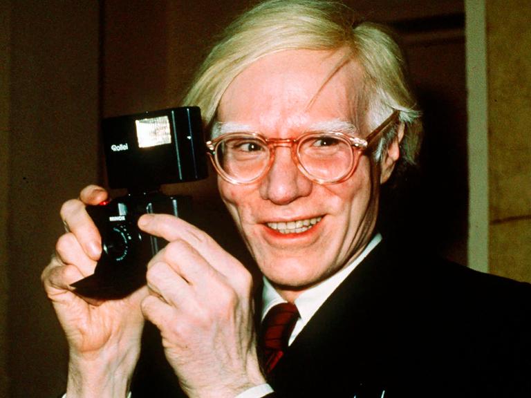 Der Künstler Andy Warhol mit einer Kamera in den Händen. (Quelle: picture alliance)