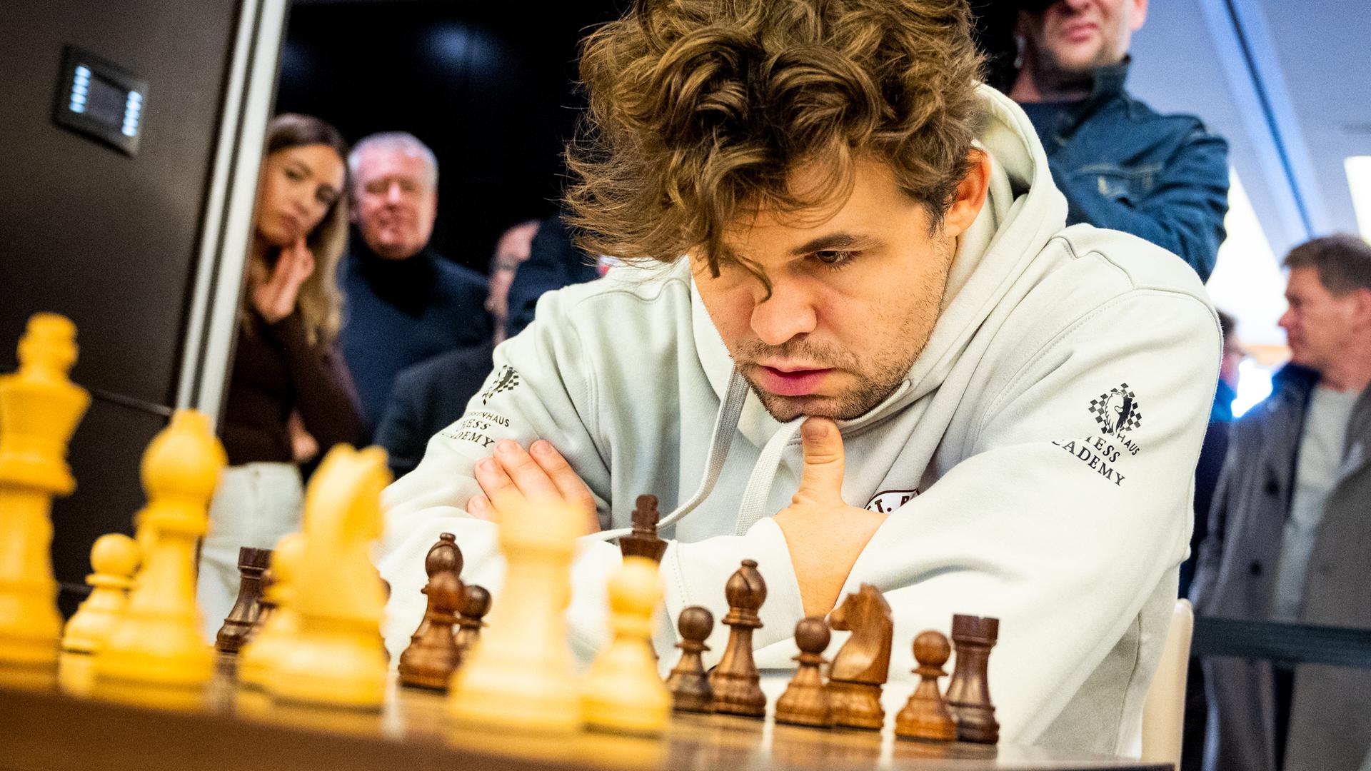 Magnus Carlsen schaut auf das Schachbrett. Er überlegt sich seinen nächsten Spielzug. Im Hintergrund stehen ein Kameramann und Publikum.