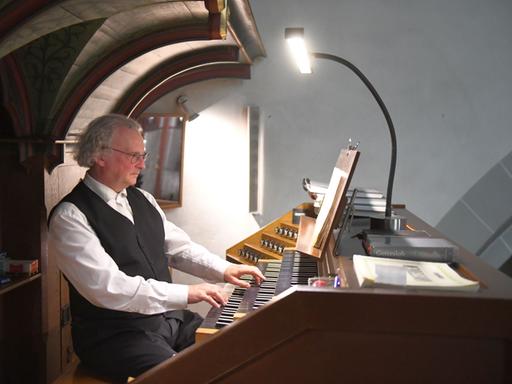 Ein Mann sitzt vor dem Spieltisch einer Orgel, die Hände sind auf den Tasten. Er trägt weißes Hemd und schwarze Weste. Er blickt konzentriert auf die Noten vor sich.