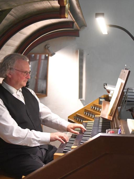 Ein Mann sitzt vor dem Spieltisch einer Orgel, die Hände sind auf den Tasten. Er trägt weißes Hemd und schwarze Weste. Er blickt konzentriert auf die Noten vor sich.