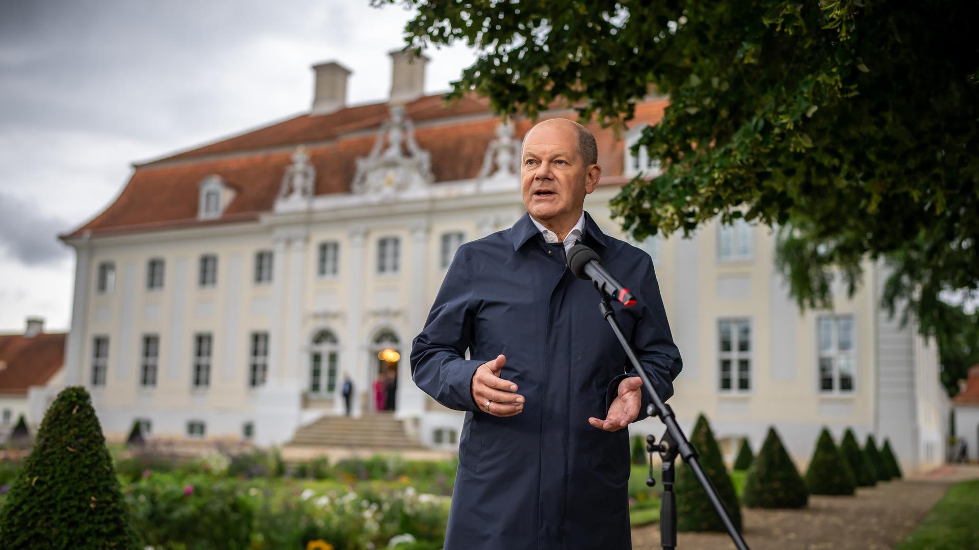 Bundeskanzler Olaf Scholz steht vor einem Mikrofon im Freien, dahinter die Fassade von Schloss Meseberg.