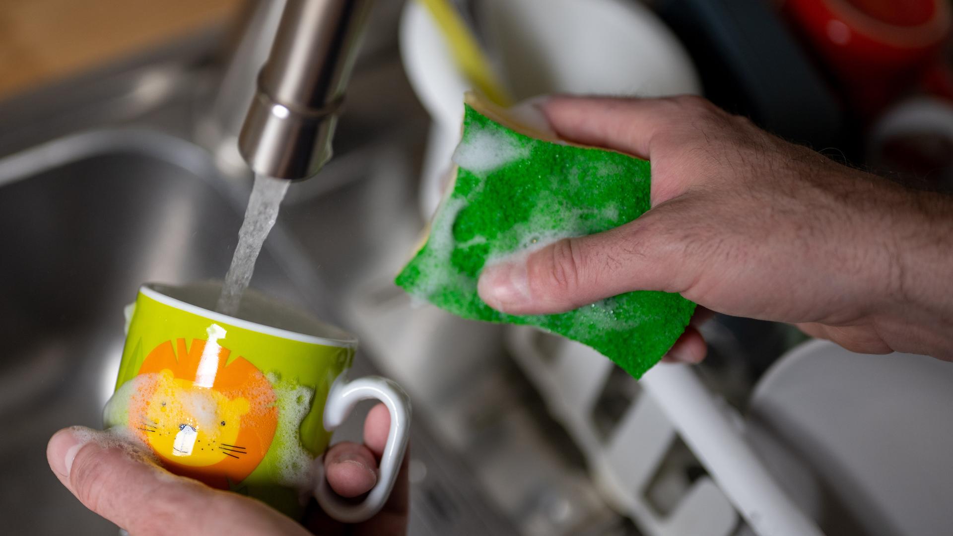 Eine Tasse wird mit einem Küchenschwamm und Spülmittel unter fließendem Wasser aus einem Wasserhahn in einer Küche gewaschen.