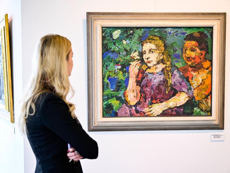 Eine blonde Frau betrachtet ein Gemälde an der Wand. Das Ölbild zeigt eine blonde Frau im rot-violetten Kleid, rechts hinter ihr ein Mann mit braunen Haaren.