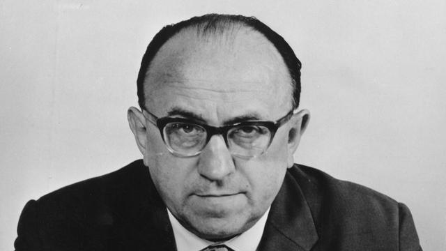Schwarz-weiße Porträtaufnahme von Alfred Müller-Armack, um 1960. Müller-Armack trägt ein weißes Hemd, Krawatte und ein dunkles Jackett und schaut durch die Gläser seiner Hornbrille in die Kamera.