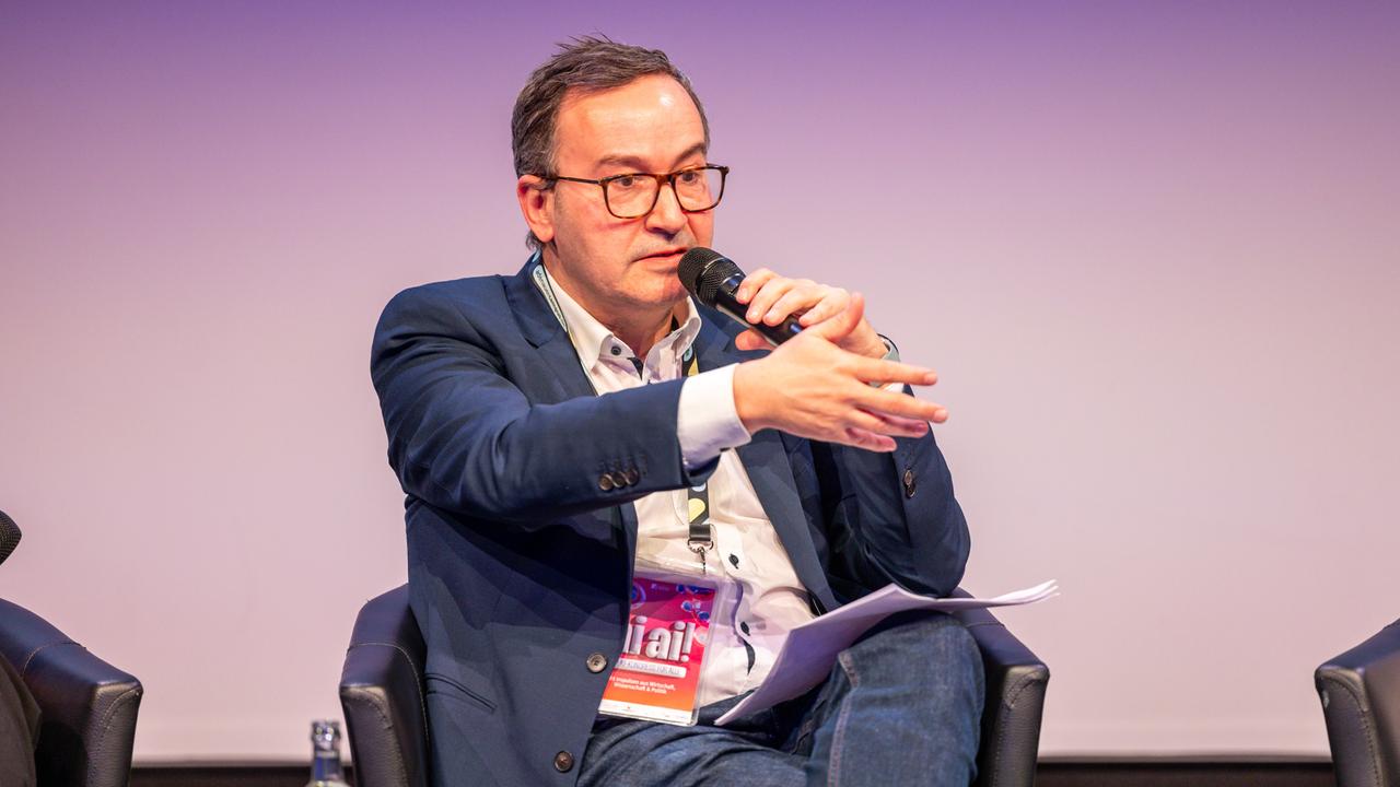 Daniel Abbou vom KI-Bundesverband sitzt bei einer Podiumsdiskussion in einem Sessel mit Mikrofon in der Hand.
