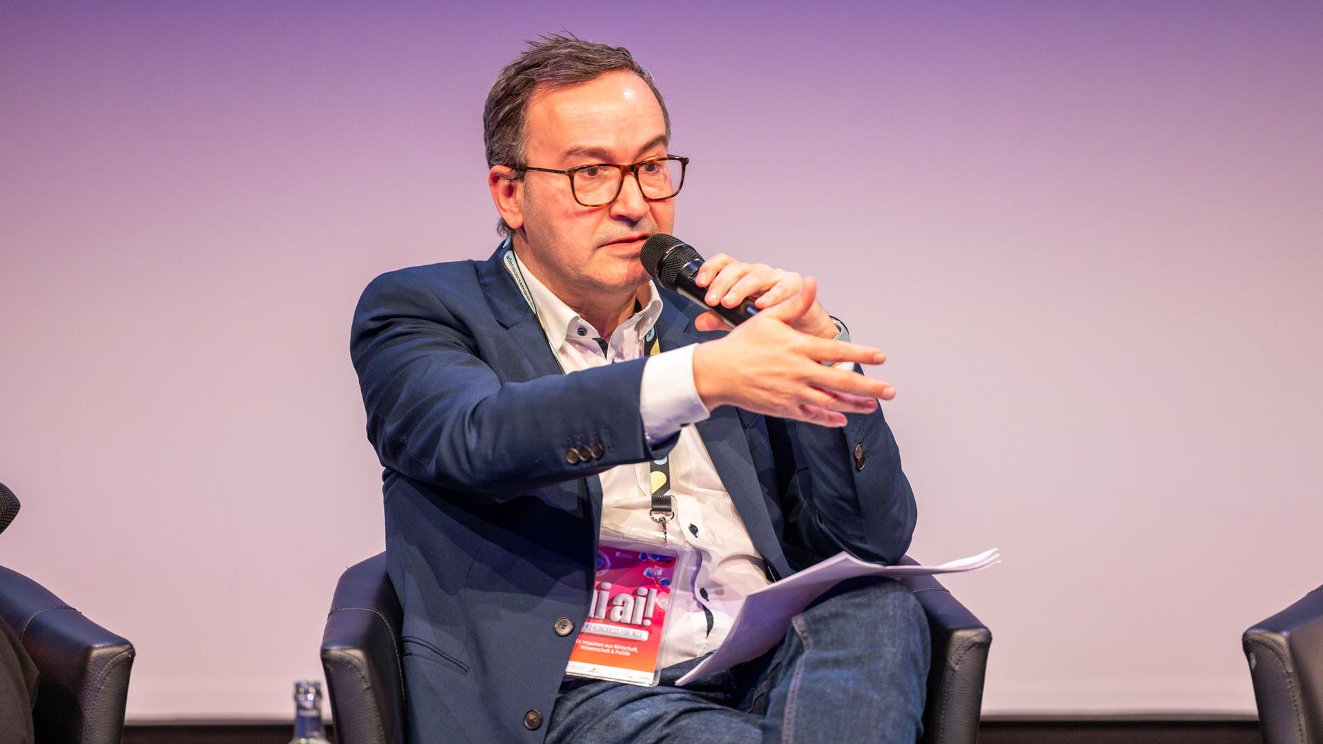 Daniel Abbou vom KI-Bundesverband sitzt bei einer Podiumsdiskussion in einem Sessel mit Mikrofon in der Hand.