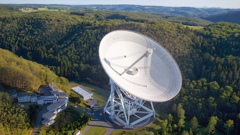 Blick auf eine weiße Radioschüssel (100 Meter Durchmesser) eines Teleskops. Rings herum ist grüner Wald.