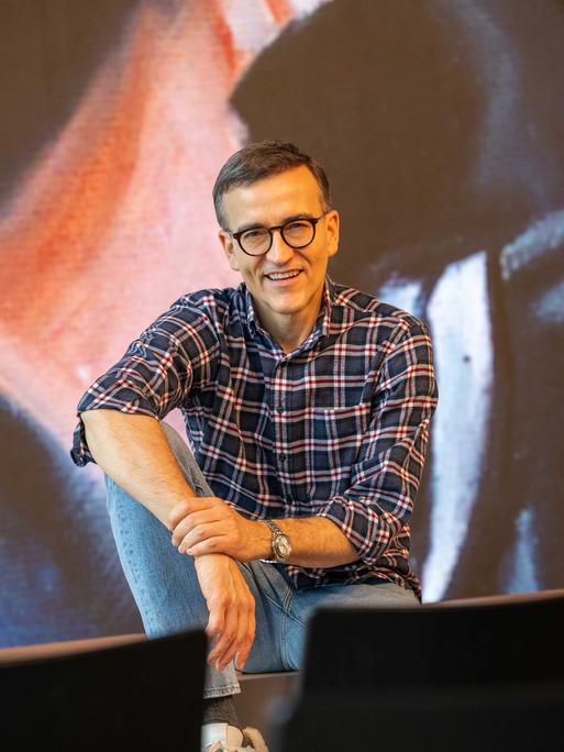 Daniel Schreiber sitzt vor einer Stuhlreihe und schaut lächelnd in die Kamera