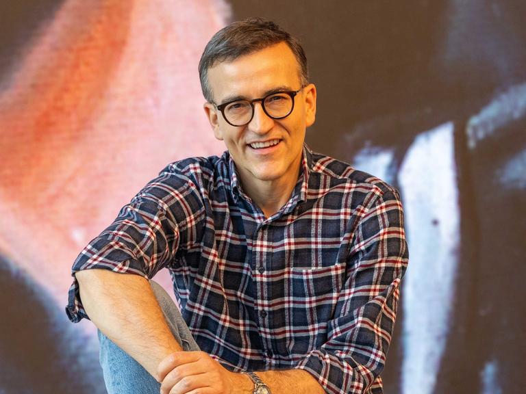 Daniel Schreiber sitzt vor einer Stuhlreihe und schaut lächelnd in die Kamera